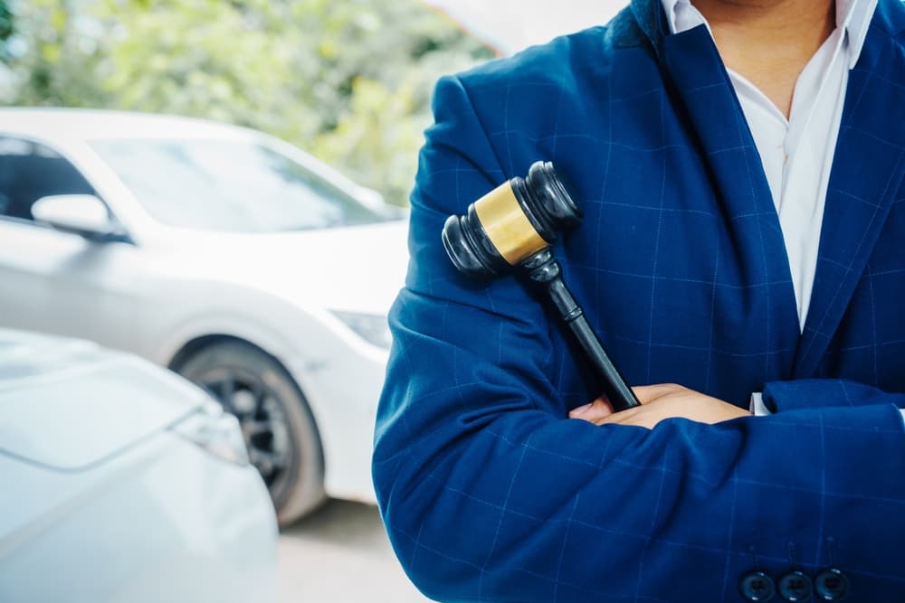 A lawyer, holding a wooden gavel, stand in front of a car, discussing legal aspects like citations, liability, and negligence related to a accident